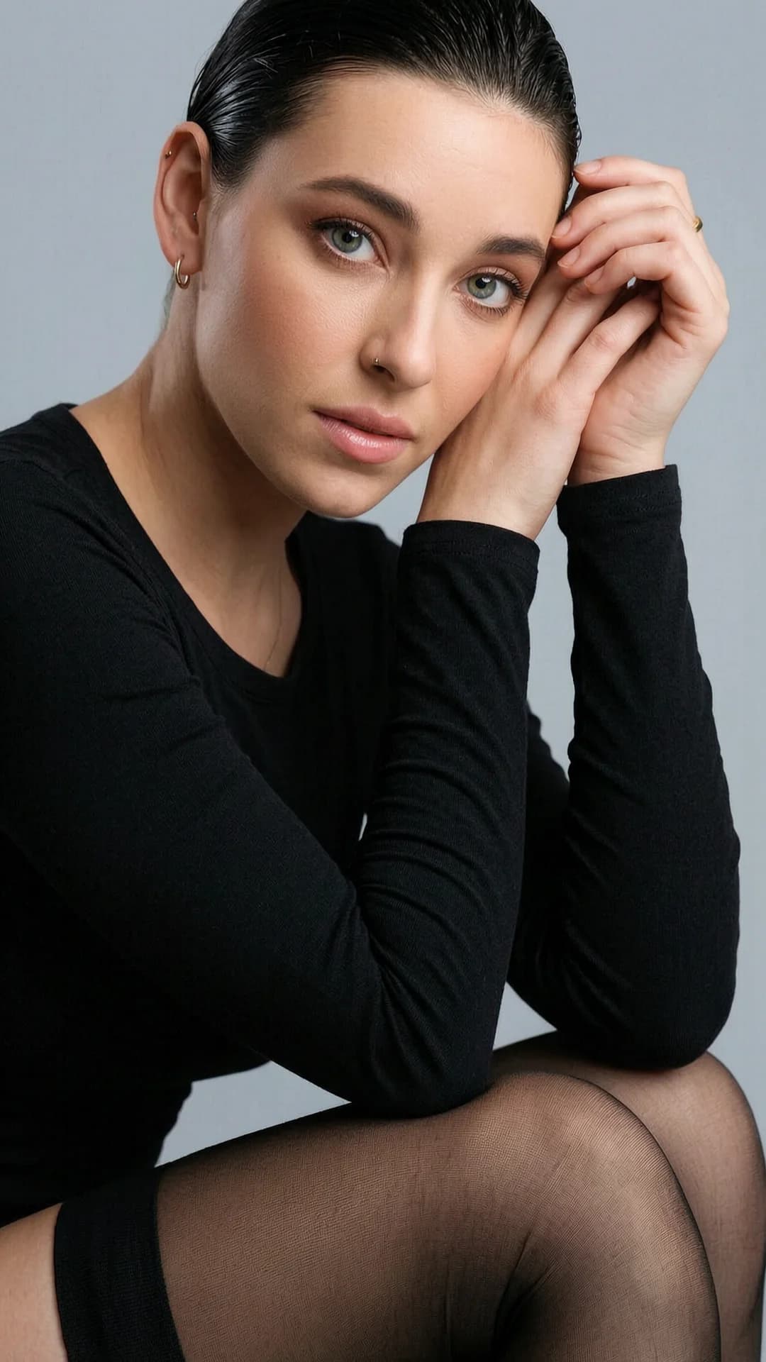 A 29-year-old Caucasian woman with a skinny body type, straight brunette hair slicked back, piercing green eyes, wearing a black long-sleeve top and sheer black stockings visible at the knee, subtle nipple piercings outlined beneath the fabric, natural makeup with clean skin and slightly glossy lips, minimal jewelry including small earrings and a gold ring, captured in a close-up portrait angled slightly to the left with shallow depth of field sharply focused on her face and softening toward the edges, her forehead resting on one hand while the other rests on her knee with fingers near her face, intense direct eye contact with the viewer conveying introspective calm and moody introspection, soft directional side lighting casting subtle natural shadows on her pale skin and plain light gray studio background, muted earth tones and black clothing, contemporary minimalistic fashion photography style, soft indirect daylight, flawless anatomy, no extra digits, no extra limbs, realistic skin texture, high-detail.