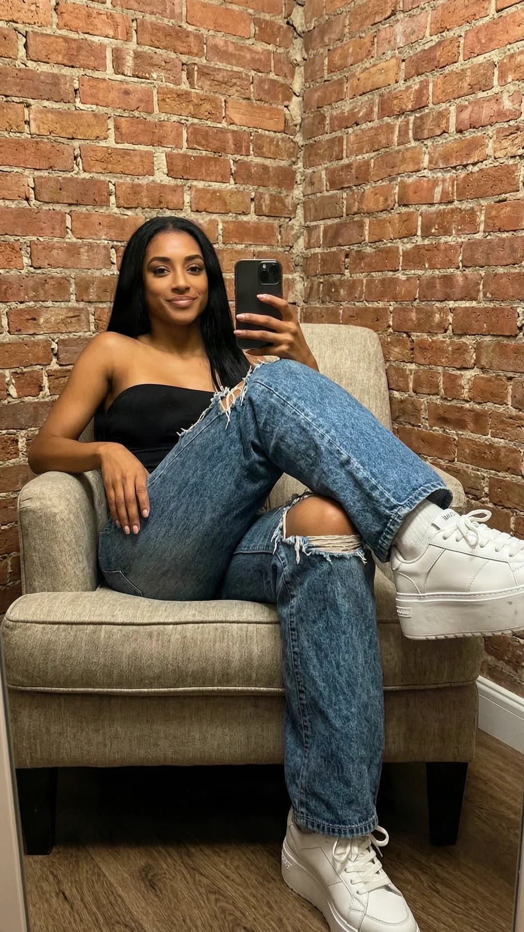 A 31-year-old skinny black woman with straight black hair, brown eyes, and small perky bare breasts visible, lounging alone in a comfortable armchair in the corner of a small dressing room with unfinished brick wall behind her, wearing only baggy ripped blue jeans and brand name white platform sneakers, captured in strict first-person selfie POV from the smartphone she is holding, her skinny torso relaxed against the chair with natural breast position and subtle skin sheen, intense direct eye contact with the camera and a wide close-lipped smile, soft warm dressing room lighting, nsfw, Flawless anatomy, no extra digits, no extra limbs, realistic skin texture, high-detail.