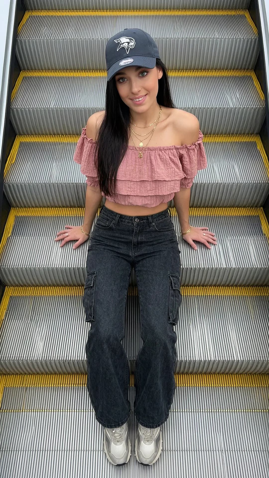 A 29-year-old Caucasian woman with skinny body type, straight brunette hair, green eyes and visible nipple piercings sits sideways on the metallic steps of a wide escalator in strict top-down vertical POV from directly above, her head resting playfully on one shoulder while making direct upward eye contact with the camera and a casual spontaneous smile, wearing a textured off-shoulder ruffled crop top in dusty pink that reveals subtle piercing outlines beneath the fabric paired with wide-leg dark denim cargo pants, chunky sneakers and layered gold necklaces, her Minnesota Vikings cap pulled low, the frame filled with rhythmic metallic ridges and yellow safety lines under natural lighting creating symmetrical urban streetwear composition, flawless anatomy, no extra digits, no extra limbs, realistic skin texture, high-detail.